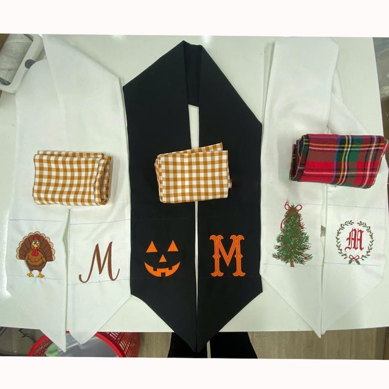 May include: Three long fabric scarves or table runners featuring seasonal embroidery. One white with a turkey and letter M, one black with a jack-o'-lantern and letter M, and one white with a Christmas tree and letter M, each with a folded fabric accent.