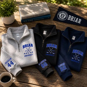 May include: A collection of Briar University quarter-zip pullovers in gray, black, and navy blue. Each pullover features the university name and crossed hockey sticks. Sleeves display names and numbers. A pennant and books are also visible.