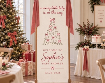 May include: A tall, rectangular fabric banner on a wooden stand, featuring the text 'a merry little baby is on the way', a line-art Christmas tree with red bows, and 'WELCOME TO Sophie's BABY SHOWER 12.25.2026'. Two large, dusty rose velvet bows adorn the top corners.