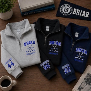 May include: Three quarter-zip sweatshirts in gray, black, and navy blue, each embroidered with "BRIAR UNIVERSITY" and crossed hockey sticks. The gray sweatshirt has "GRAHAM 44" on the sleeve, black has "DI LAURENTIS 66", and navy has "LOGAN 22". A pennant with "BRIAR UNIVERSITY" is also visible.