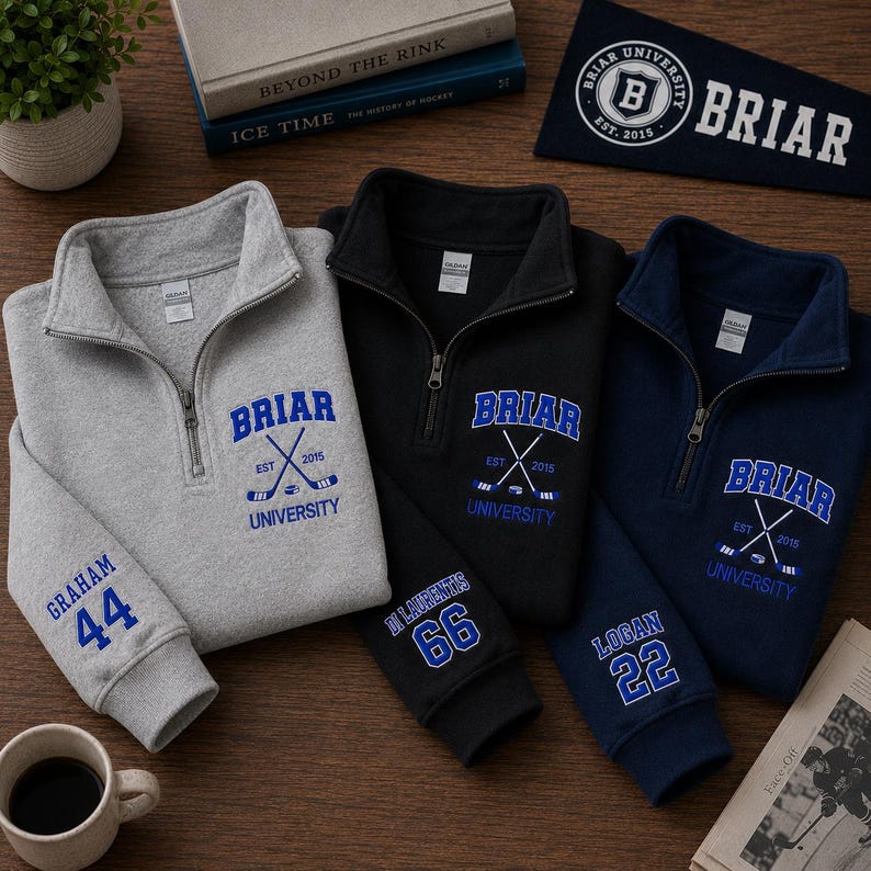 May include: Three quarter-zip sweatshirts in gray, black, and navy blue, each embroidered with "BRIAR UNIVERSITY" and crossed hockey sticks. The gray sweatshirt has "GRAHAM 44" on the sleeve, black has "DI LAURENTIS 66", and navy has "LOGAN 22". A pennant with "BRIAR UNIVERSITY" is also visible.