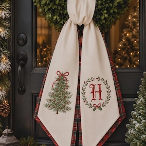 May include: A cream-colored fabric door hanger featuring a red and green embroidered Christmas tree on one side and a red letter H inside a green wreath on the other. The fabric has a red and green plaid backing and is tied in a knot at the top.