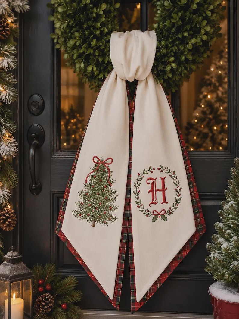 May include: A cream-colored fabric door hanger featuring a red and green embroidered Christmas tree on one side and a red letter H inside a green wreath on the other. The fabric has a red and green plaid backing and is tied in a knot at the top.