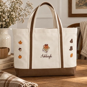 Könnte beinhalten: Eine cremefarbene Canvas-Tragetasche mit braunen Rändern und Griffen. Die Vorderseite zeigt gestickte Herbstmotive wie ein Ahornblatt, eine Eichel, einen Kürbis, eine Mütze, Stiefel und ein Eichenblatt. Mittig sind Bücher mit Tasse und der Name Ashleigh.