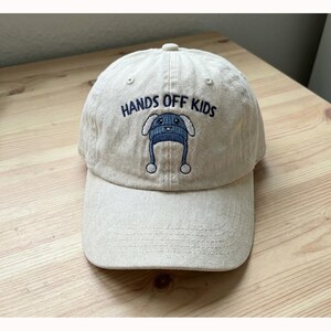 May include: A beige baseball cap with the text "HANDS OFF KIDS" embroidered in black. The cap features an embroidered design of a blue hat with ear flaps. The cap has a curved brim and a button on top.