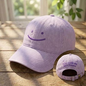 May include: A lilac baseball cap featuring an embroidered smiling face on the front. The back of the cap has the embroidered text "YOU CAN BE ANYTHING". The cap has an adjustable strap.
