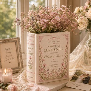 May include: A rectangular book-shaped vase in cream with pink floral illustrations and text reading Our Love Story, A Beautiful Love Story, Written By Olivia & James, 06.14.2025, and Every chapter is better together, filled with pink flowers.