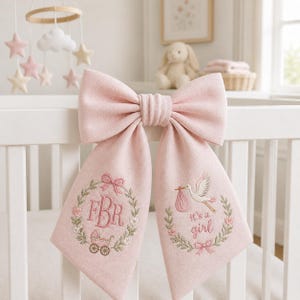 Könnte beinhalten: Eine große rosa Schleife mit gestickten Details, die an einem weißen Kinderbett hängt. Die Schleife zeigt einen Blumenkranz mit den Initialen "FBR" und einen Kinderwagen. Die andere Seite hat einen Storch und den Text "it's a girl".