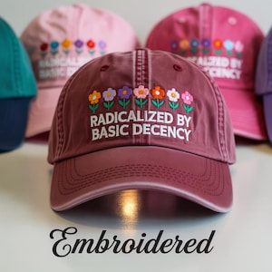 Op de afbeelding: Een marineblauwe baseballpet met geborduurde bloemen en de tekst "Radicalized by Basic Decency". De pet is gemaakt van zacht, comfortabel materiaal en heeft een gebogen klep.