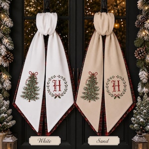 May include: Two hanging decorative towels, one white and one sand-colored, each featuring a red embroidered letter H inside a green wreath and a green Christmas tree with a red bow. The reverse side of both towels displays a red and green plaid pattern.