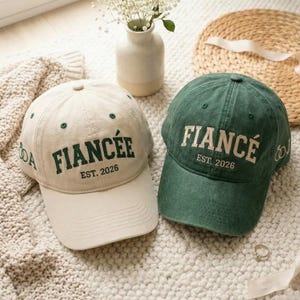 Puede incluir: Dos gorras de béisbol, una beige y otra verde, con las palabras "FIANCÉE" y "FIANCÉ" bordadas en verde y blanco, respectivamente. Ambas gorras tienen "EST. 2026" debajo del texto principal y un pequeño gráfico de anillo. Artículos de boda.