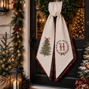 Peut inclure: Un accessoire de porte en tissu crème drapé sur une couronne verte, avec un envers à carreaux rouges et verts. Le panneau gauche présente un sapin de Noël décoré d'un nœud rouge, tandis que le panneau droit montre une lettre H rouge dans une couronne.