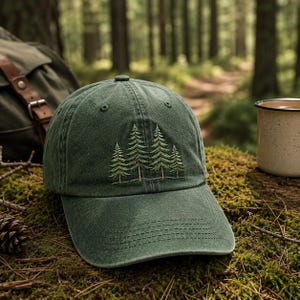 May include: A forest green baseball cap featuring an embroidered design of four pine trees in varying heights. The cap has a structured crown, a curved brim with tonal stitching, and a button at the top center. The fabric has a washed texture.