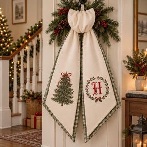 May include: A cream-colored fabric door hanger featuring a large bow at the top, accented with a pine and berry wreath. One panel displays an embroidered green Christmas tree with a red bow, while the other shows a red letter H inside a green wreath.