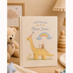 May include: A cream-colored fabric-covered memory book featuring embroidered designs of a yellow dinosaur wearing a party hat, a small rabbit, a teddy bear, and a rainbow. The text reads Little Moments of Oliver James in blue script.