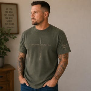 May include: An olive green short-sleeved t-shirt featuring a thin vertical cross graphic on the chest with the text "FATHERHOOD IS KINGDOM WORK" and "PROVERBS 22:6". The left sleeve displays the text "Hann Isaac" with a small cross symbol.