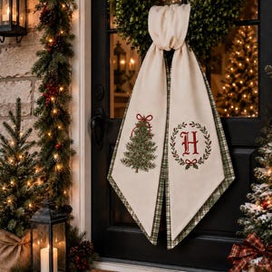 May include: A cream-colored fabric door hanger featuring a green Christmas tree with a red bow on one side and a red letter H inside a green wreath on the other. The fabric has a green and white plaid backing and hangs from a green wreath.