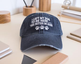 May include: A dark blue denim baseball cap with white embroidered text that reads "I left my dog to be here... this better be good" and four white paw prints.