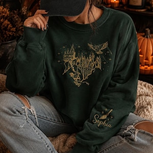 Puede incluir: Una sudadera de cuello redondo verde oscuro con una ilustración de líneas doradas de un castillo y un búho en el pecho. La manga izquierda muestra un gráfico de una escoba dorada sobre el nombre Sophia bordado en letra cursiva dorada.