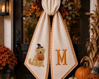 May include: A cream-colored fabric door hanging featuring an embroidered golden retriever wearing a witch hat and holding a pumpkin bucket on one side, and a large orange letter M on the other, both bordered by an orange and white gingham trim.