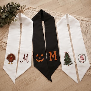 May include: Three long, pointed fabric sashes. The left is white with an embroidered turkey and the letter M. The center is black with an orange jack-o'-lantern and the letter M. The right is white with an embroidered Christmas tree and a wreath.