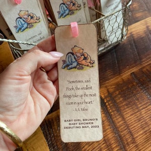 K&ouml;nnte beinhalten: Ein Winnie Puuh und Ferkel Lesezeichen mit einem Zitat von A.A. Milne. Das Lesezeichen ist beige mit einem rosa Band. Der Text lautet "Sometimes, said Pooh, the smallest things take up the most room in your heart." -A.A. Milne. Baby Girl Bruno's Baby Shower Debuting May, 2023.