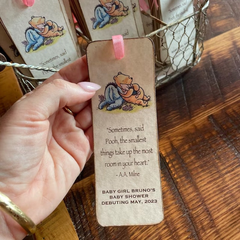 K&ouml;nnte beinhalten: Ein Winnie Puuh und Ferkel Lesezeichen mit einem Zitat von A.A. Milne. Das Lesezeichen ist beige mit einem rosa Band. Der Text lautet "Sometimes, said Pooh, the smallest things take up the most room in your heart." -A.A. Milne. Baby Girl Bruno's Baby Shower Debuting May, 2023.