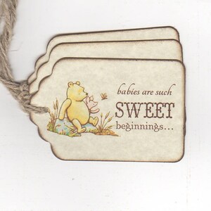 May include: A Winnie the Pooh themed baby shower favor tag with a brown string. The tag is beige with brown edges and features a cartoon image of Winnie the Pooh and Piglet. The text on the tag reads "babies are such SWEET beginnings..."