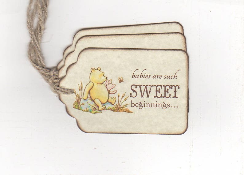 May include: A Winnie the Pooh themed baby shower favor tag with a brown string. The tag is beige with brown edges and features a cartoon image of Winnie the Pooh and Piglet. The text on the tag reads "babies are such SWEET beginnings..."