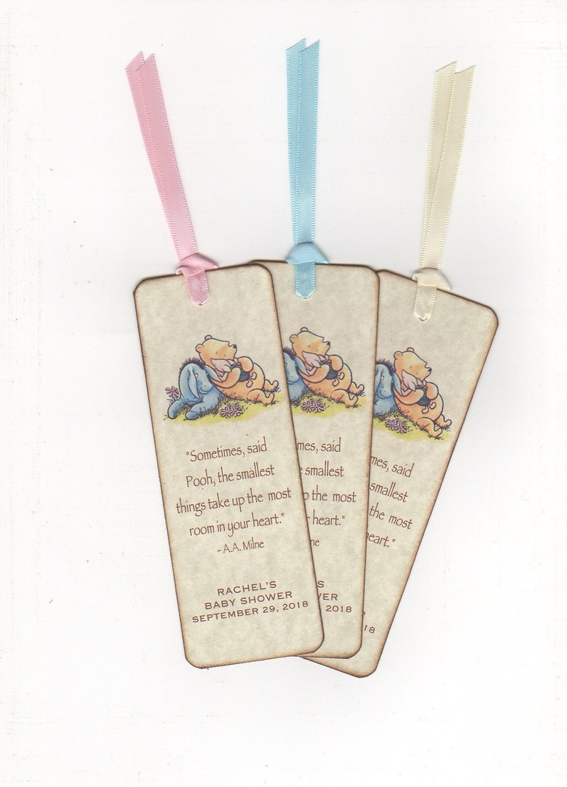 K&ouml;nnte beinhalten: Drei Winnie Puuh Lesezeichen-Geschenke mit rosa, blauem und cremefarbenem Band. Die Lesezeichen zeigen ein Zitat von A.A. Milne: "Manchmal, sagte Winnie Puuh, nehmen die kleinsten Dinge den meisten Platz in deinem Herzen ein." Die Lesezeichen sind f&uuml;r Rachels Babyparty am 29. September 2018.