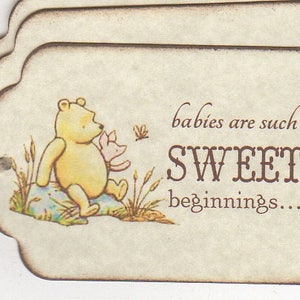 May include: A Winnie the Pooh themed baby shower favor tag with a brown background and a picture of Winnie the Pooh and Piglet. The text on the tag reads "babies are such SWEET beginnings..."