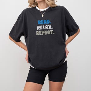 May include: Black t-shirt with the words "READ. RELAX. REPEAT." in blue and white text. The shirt is worn with black shorts and a white undershirt. The model is wearing a necklace.
