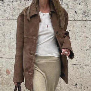 May include: A brown suede jacket with a button-up front, worn over a white ribbed top and tan skirt. The person is wearing sunglasses and carrying a brown handbag. The background is a textured stone wall.