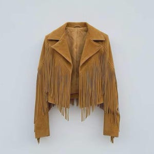 May include: A tan suede jacket with long fringe detailing on the sleeves and chest. The jacket has a classic collar and a tailored fit, perfect for a bohemian or western-inspired look. The jacket is displayed against a plain white background.