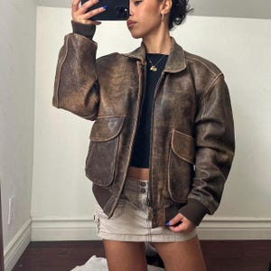 Può includere: Giacca bomber in pelle marrone con finitura invecchiata. La giacca presenta una chiusura con zip, due tasche frontali e polsini e orlo a coste. Ha un colletto classico e un'estetica vintage.