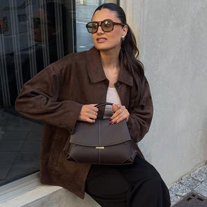 May include: A woman wearing a brown suede jacket, sunglasses, and holding a dark brown handbag. The handbag has a structured design with a gold clasp. She is also wearing a white top and black pants.