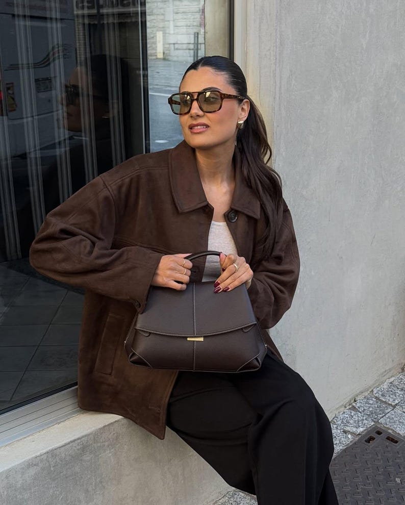 May include: A woman wearing a brown suede jacket, sunglasses, and holding a dark brown handbag. The handbag has a structured design with a gold clasp. She is also wearing a white top and black pants.