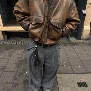 May include: A brown leather bomber jacket with a zip fastening and two large pockets. The jacket is worn over a grey pair of trousers with a black belt and a silver chain.