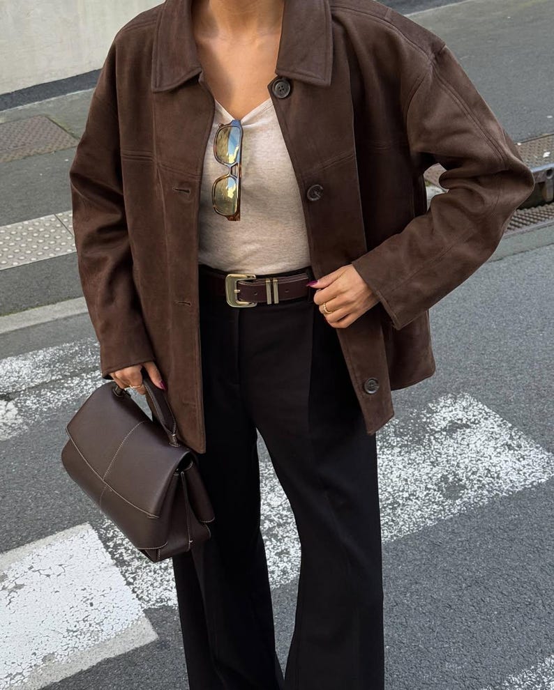 May include: A brown suede jacket with a collared neckline and button closure. The jacket is paired with a light-colored top, black pants, and a brown leather belt. A brown handbag is held in the hand.