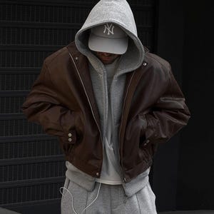 Puede incluir: Chaqueta bomber de cuero marrón con una sudadera con capucha gris debajo. La persona lleva una gorra de béisbol gris con el logo "NY" blanco y pantalones de chándal grises. La chaqueta tiene cierre de cremallera y botones.