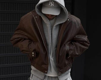 May include: A brown leather bomber jacket with a gray hooded sweatshirt underneath. The person is wearing a gray baseball cap with a white "NY" logo and gray sweatpants. The jacket has a zipper closure and button details.