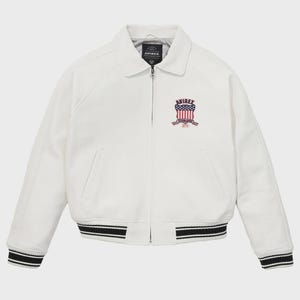 K&ouml;nnte beinhalten: Eine wei&szlig;e Jacke mit Kragen und Rei&szlig;verschluss. Die Jacke hat einen Aufn&auml;her mit dem Text "AVIREX" und einem amerikanischen Flaggen-Design. Die Manschetten und der Saum haben schwarz-wei&szlig;e Streifen. Ein klassisches Oberbekleidungsst&uuml;ck.