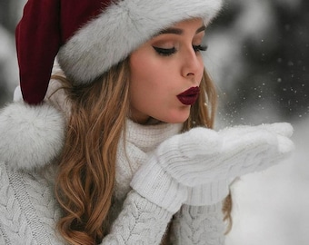 May include: A person wearing a burgundy Santa hat with white faux fur trim, a white cable-knit jumper, and white mittens. She is blowing snow, with a snowy background.