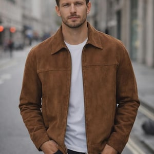 May include: A brown suede jacket with a classic collar, a full-length front zipper, and long sleeves, worn over a plain white t-shirt and dark blue denim jeans. The jacket features a horizontal seam across the chest and two side pockets.
