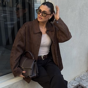 May include: A brown suede jacket with a collared neckline and a matching brown handbag. The handbag has a structured design with a top handle and a rectangular shape. The person is wearing sunglasses and black pants.