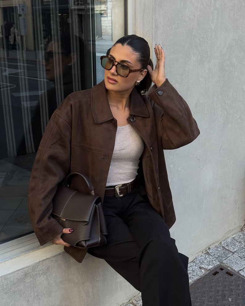 May include: A brown suede jacket with a collared neckline and a matching brown handbag. The handbag has a structured design with a top handle and a rectangular shape. The person is wearing sunglasses and black pants.