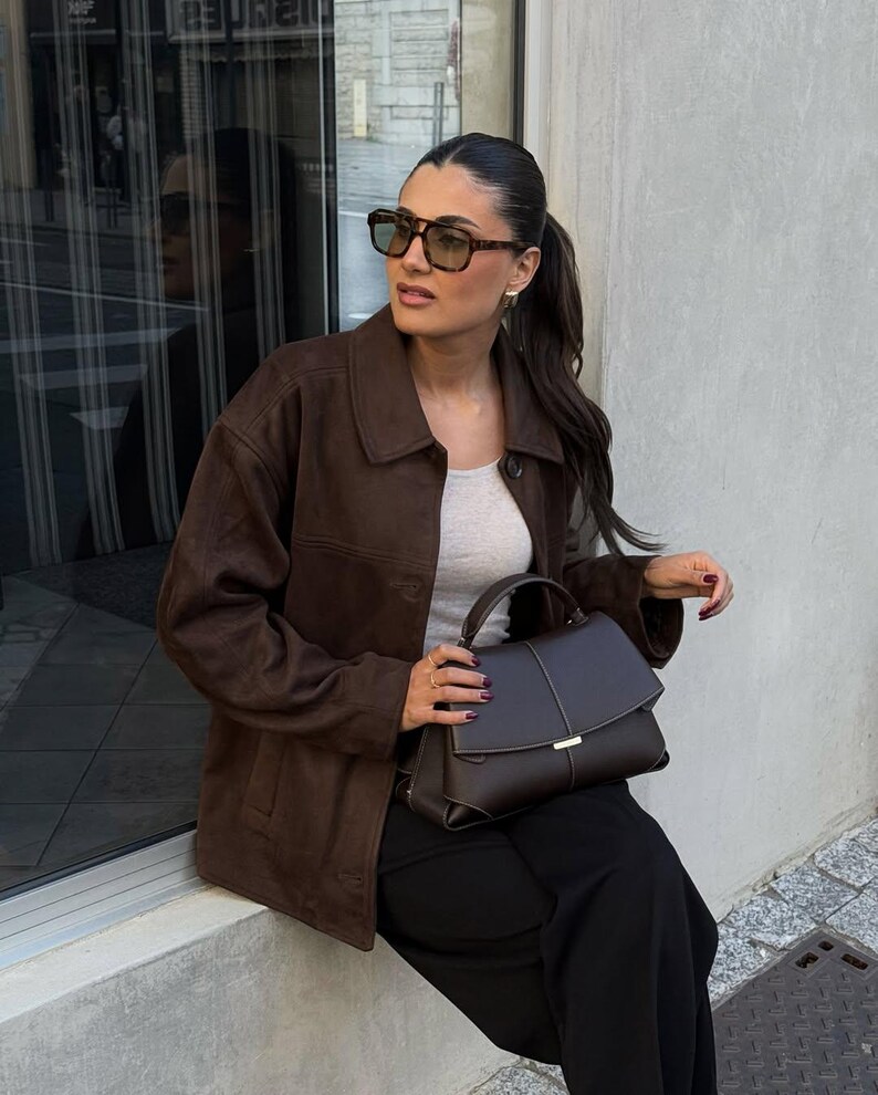 May include: A woman wearing brown sunglasses, a brown suede jacket, a gray top, and black pants. She is holding a dark brown handbag with a gold clasp. The background includes a window and a concrete wall.