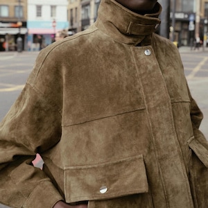 Puede incluir: Chaqueta de ante marrón con cuello alto y dos bolsillos delanteros. La chaqueta tiene cierre de botón y un corte relajado. La tela tiene un aspecto suave y texturizado. La chaqueta es de color neutro, adecuada para varios conjuntos.