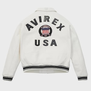 K&ouml;nnte beinhalten: Eine wei&szlig;e Jacke mit schwarz-wei&szlig; gestreiften B&uuml;ndchen und Saum. Auf der R&uuml;ckseite steht das Wort "AVIREX" &uuml;ber einem runden Aufn&auml;her mit US-Flaggen-Design und den Worten "ALL AMERICANS ESTABLISHED 1975". Darunter steht das Wort "USA".