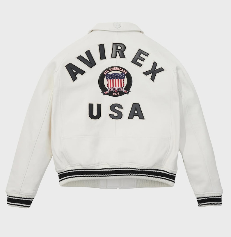 K&ouml;nnte beinhalten: Eine wei&szlig;e Jacke mit schwarz-wei&szlig; gestreiften B&uuml;ndchen und Saum. Auf der R&uuml;ckseite steht das Wort "AVIREX" &uuml;ber einem runden Aufn&auml;her mit US-Flaggen-Design und den Worten "ALL AMERICANS ESTABLISHED 1975". Darunter steht das Wort "USA".